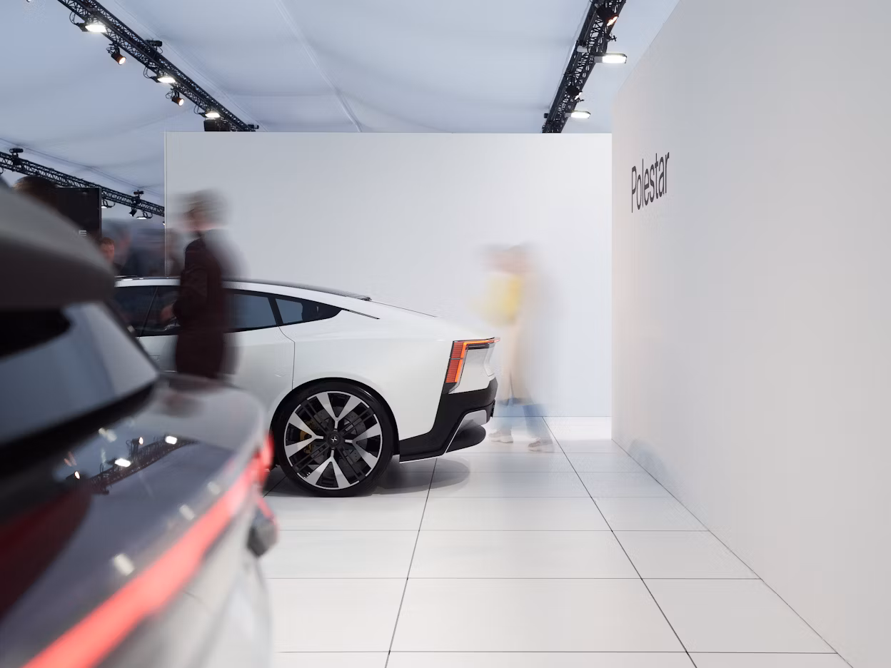 White Polestar car displayed indoors with people walking nearby, Polestar branding visible on the wall.
