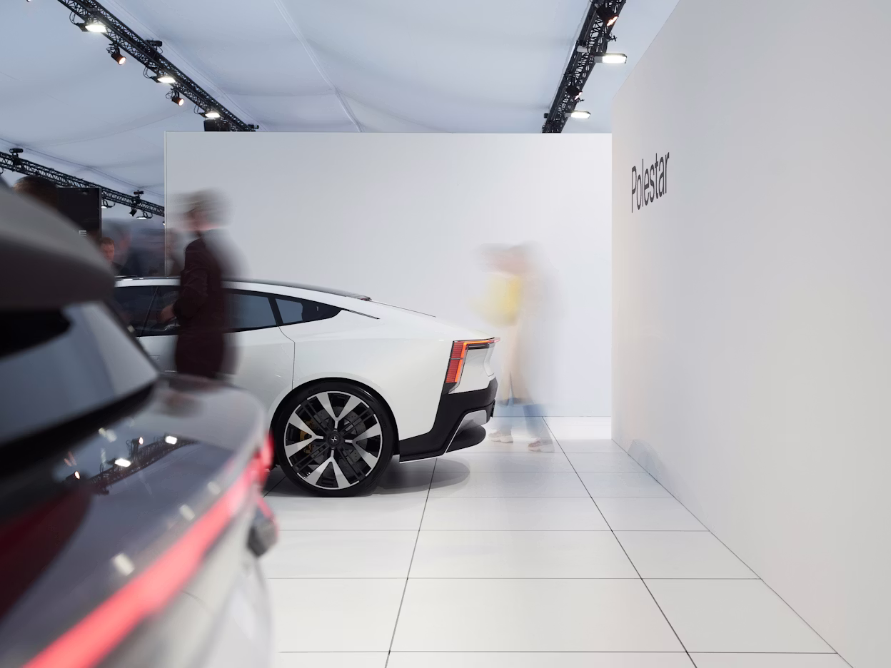 White Polestar car displayed indoors with people walking nearby, Polestar branding visible on the wall.