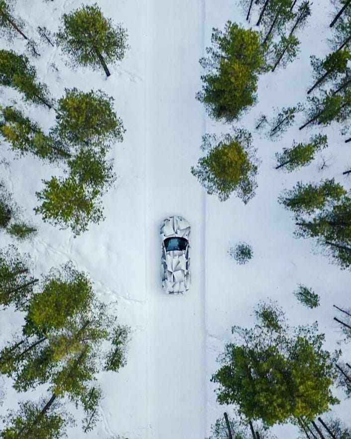 Brid's-eye view of a car driving on a snowy road surrounded by tall pine trees.
