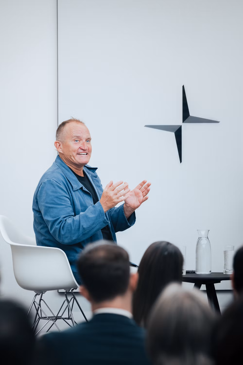 Person seated on a white chair gesturing with hands, Polestar logo on wall, table with glassware in foreground.