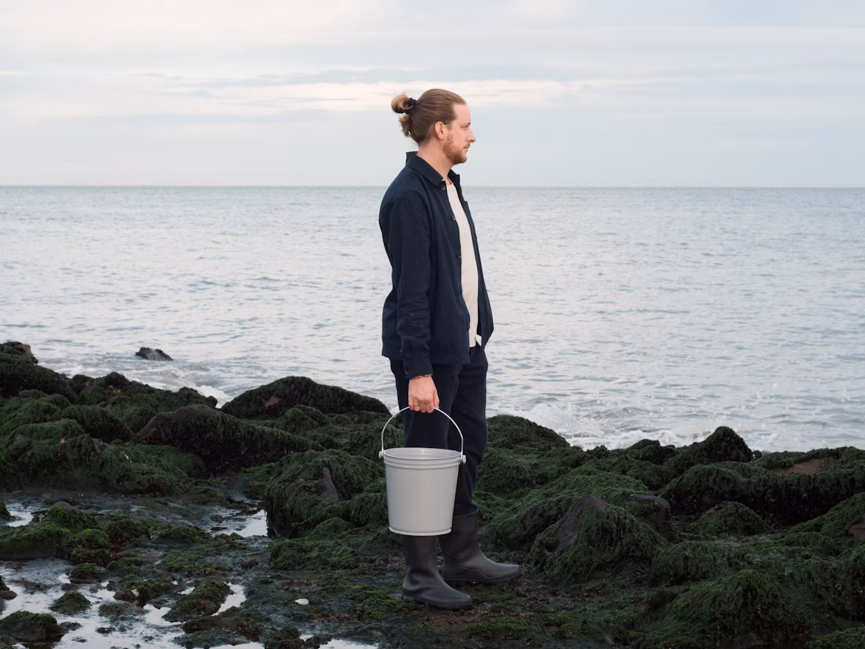 Person standing on moss-covered rocks near the sea holding a bucket.