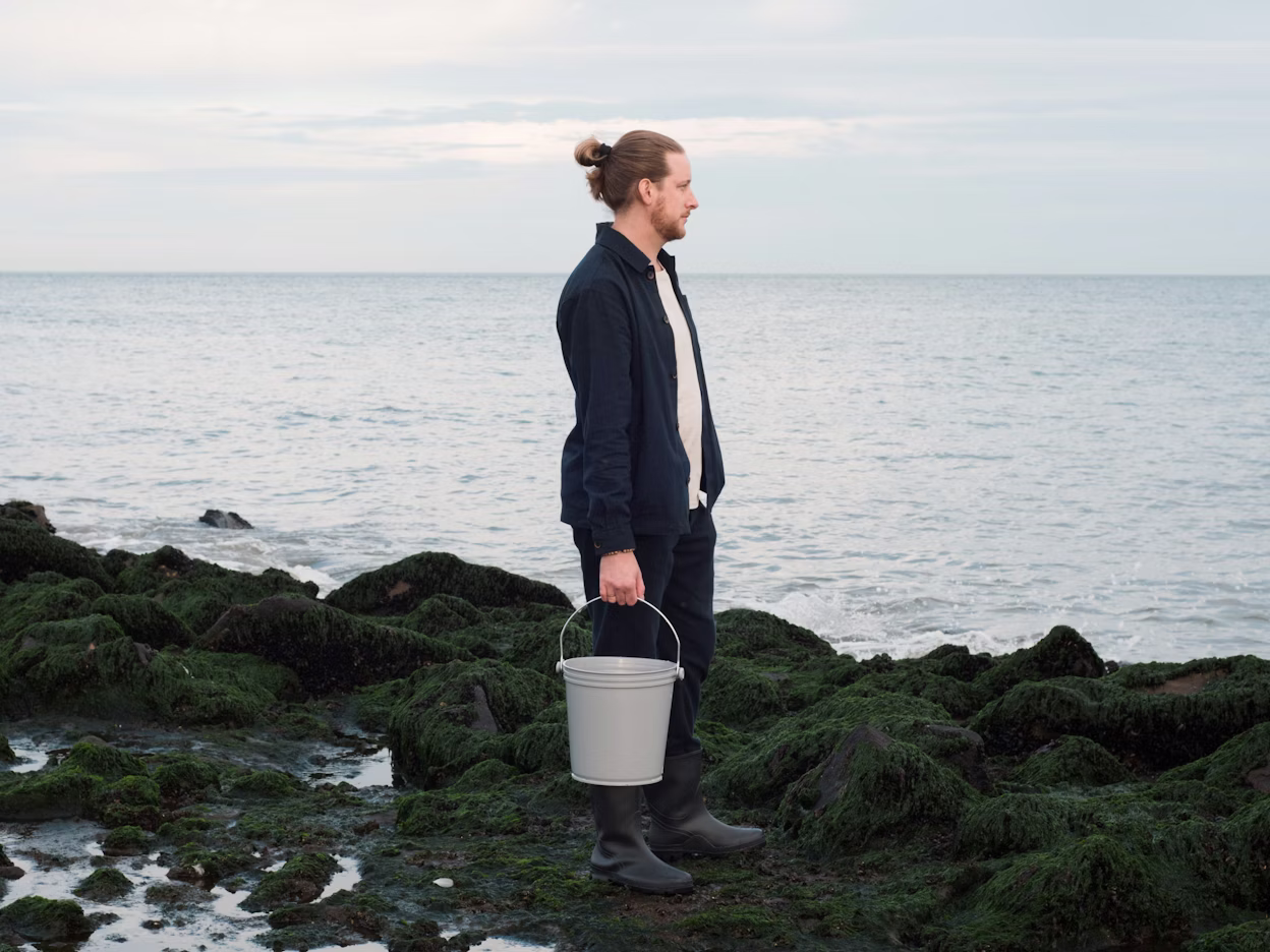 Person standing on moss-covered rocks near the sea holding a bucket.