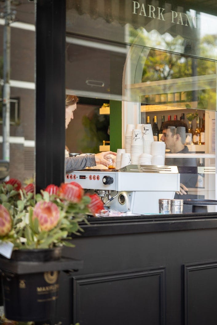 A photograph of a barista making coffee, seen through a window.