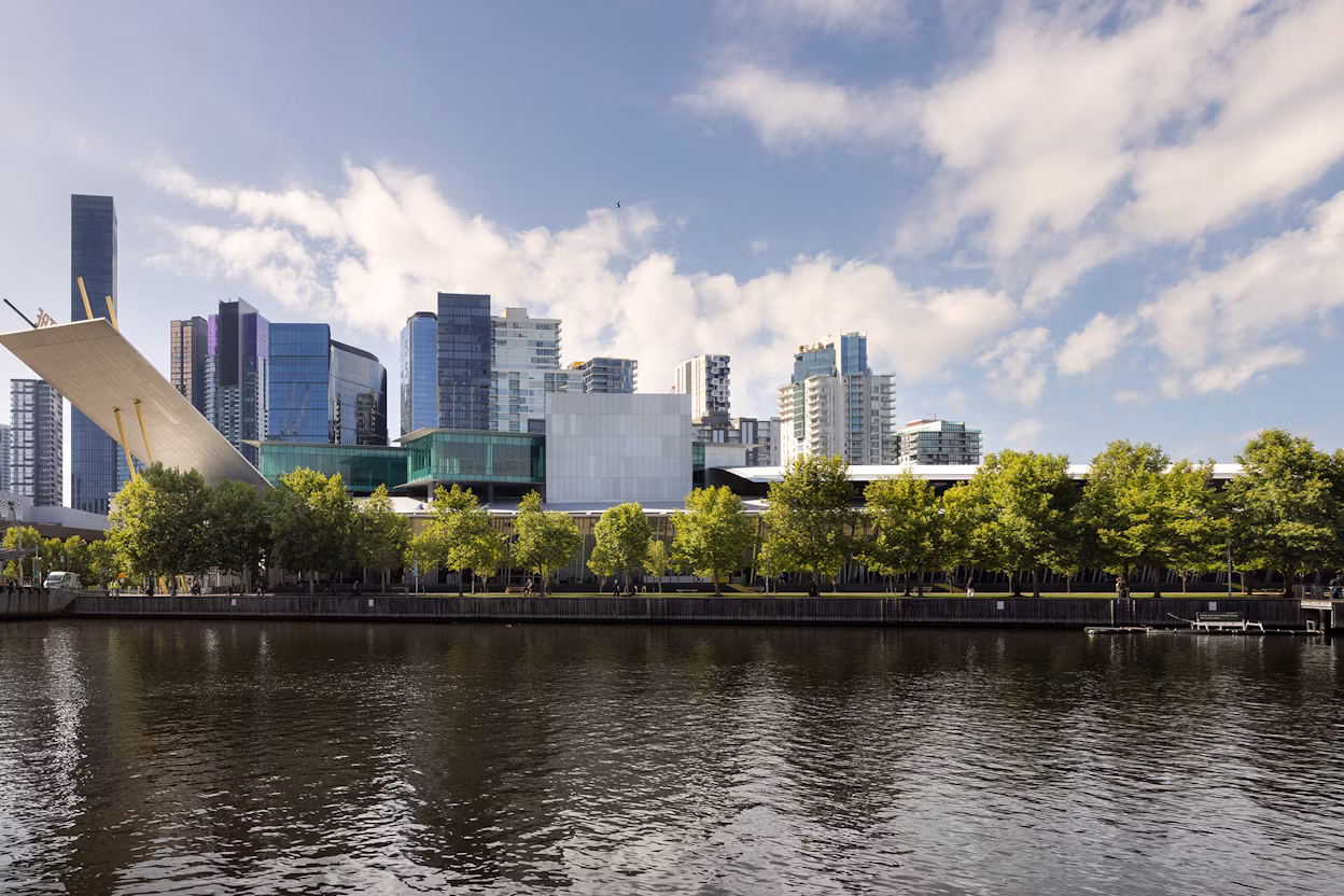 City skyline with modern buildings, trees, and a river in the foreground under a partly cloudy sky.