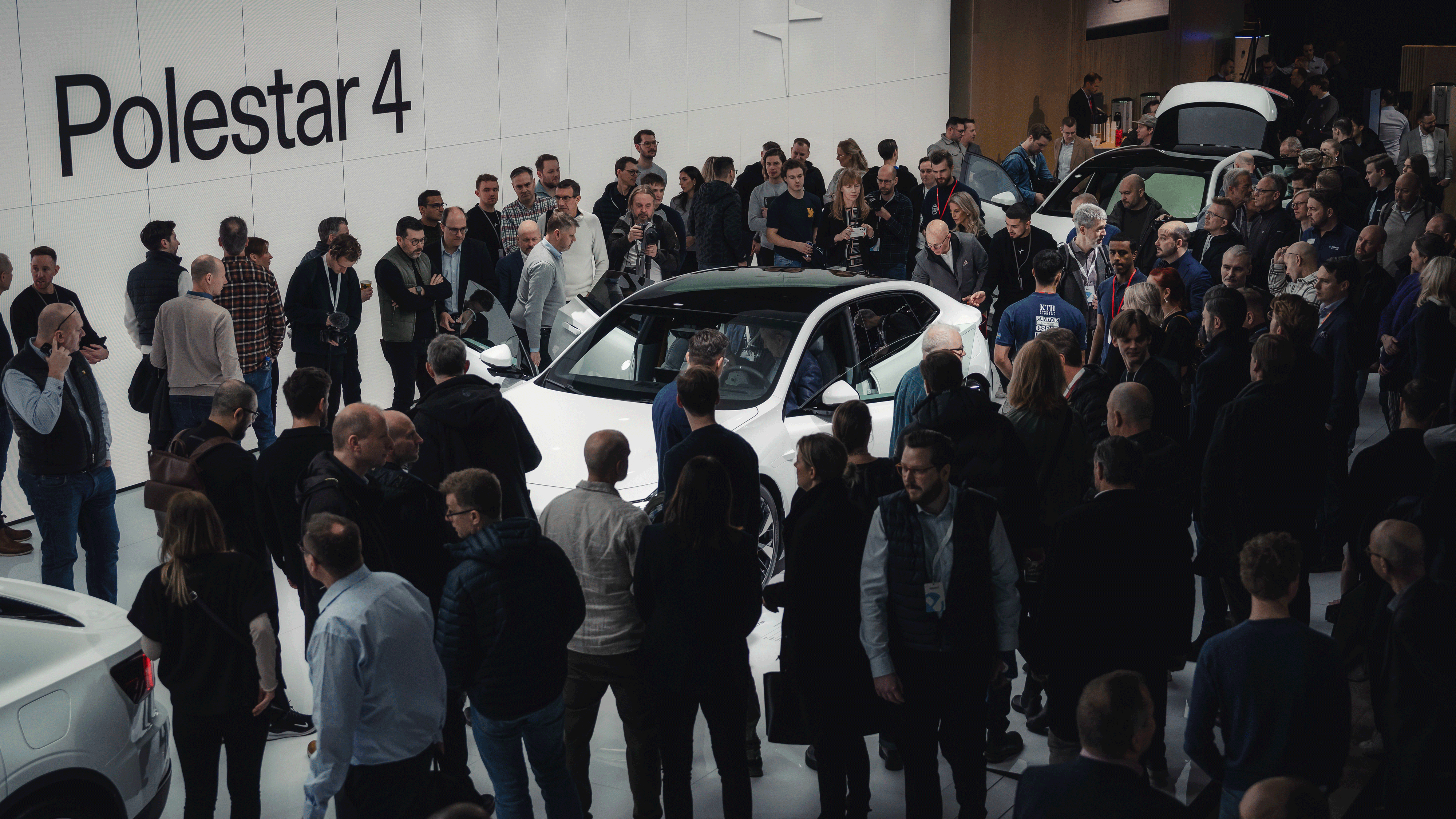 Crowd gathered around a white Polestar 4 car in an indoor event space with Polestar branding on the wall.