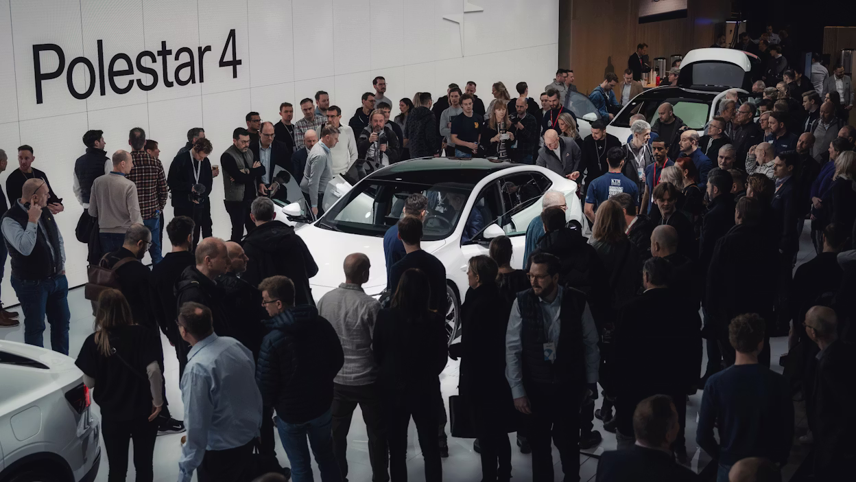 Crowd gathered around a white Polestar 4 car in an indoor event space with Polestar branding on the wall.
