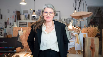 Person in formal attire standing in a workspace with design materials, wooden objects, and hanging lights visible.
