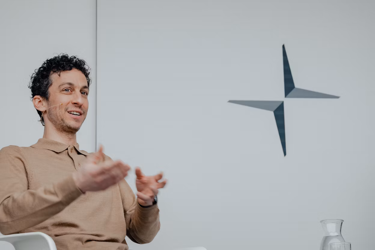 Logo of Polestar on a wall, person gesturing with hands, glass carafe on table.