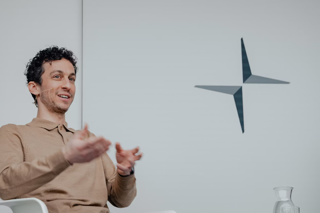 Logo of Polestar on a wall, person gesturing with hands, glass carafe on table.