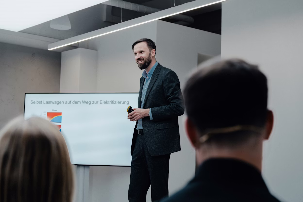 Person presenting in a room with a screen displaying text about truck electrification, audience visible in foreground.