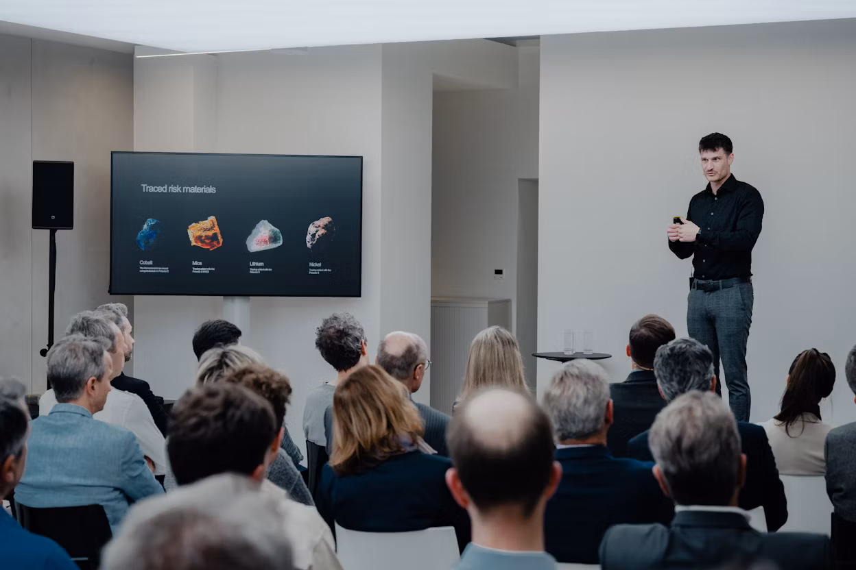 Speaker presenting traced risk materials cobalt, mica, lithium, nickel on screen to seated audience in modern room.