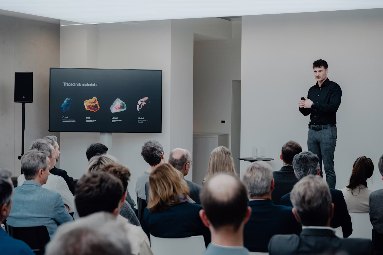 Speaker presenting traced risk materials cobalt, mica, lithium, nickel on screen to seated audience in modern room.