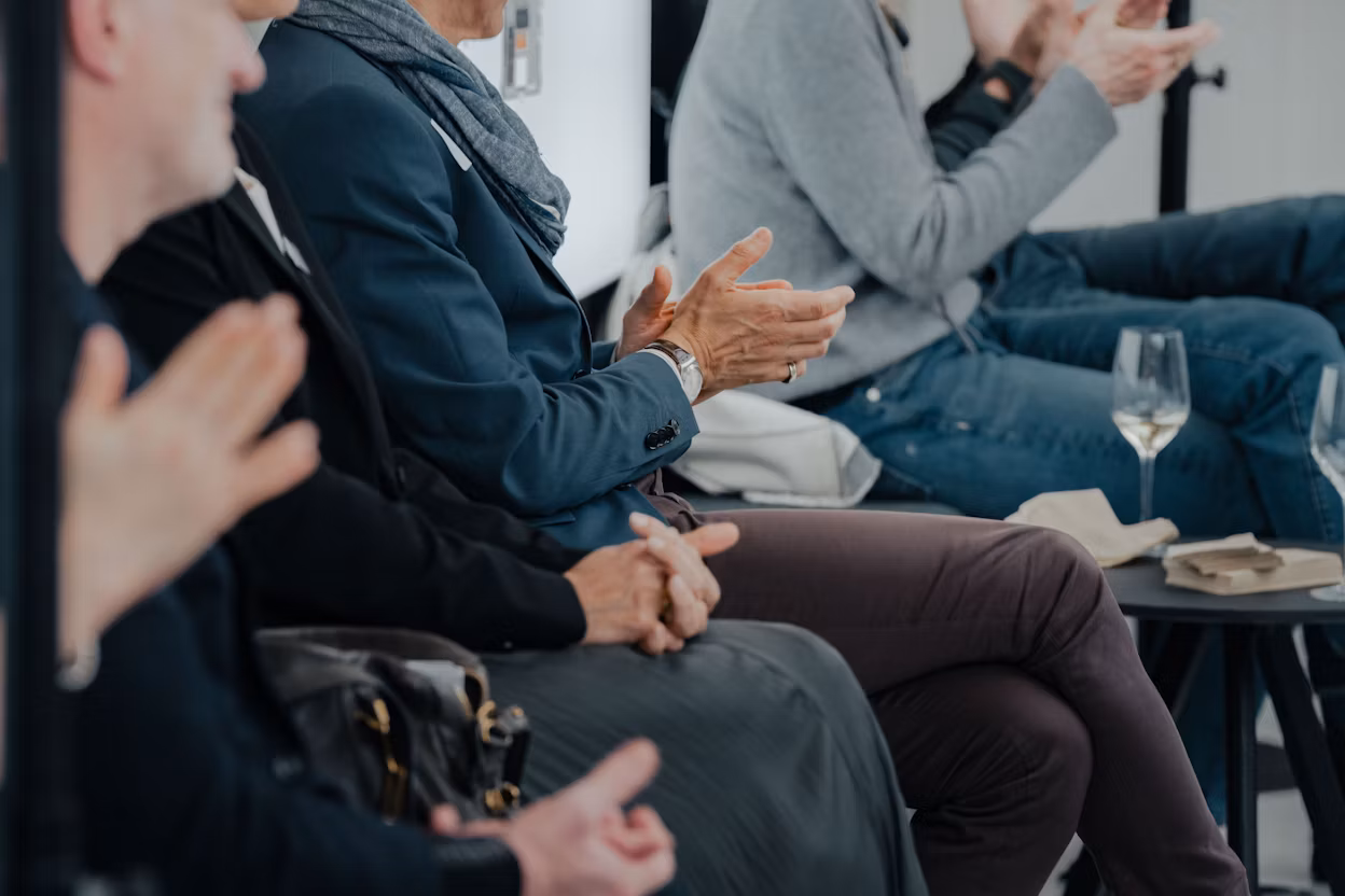 People seated clapping, with glasses of wine on a table nearby.