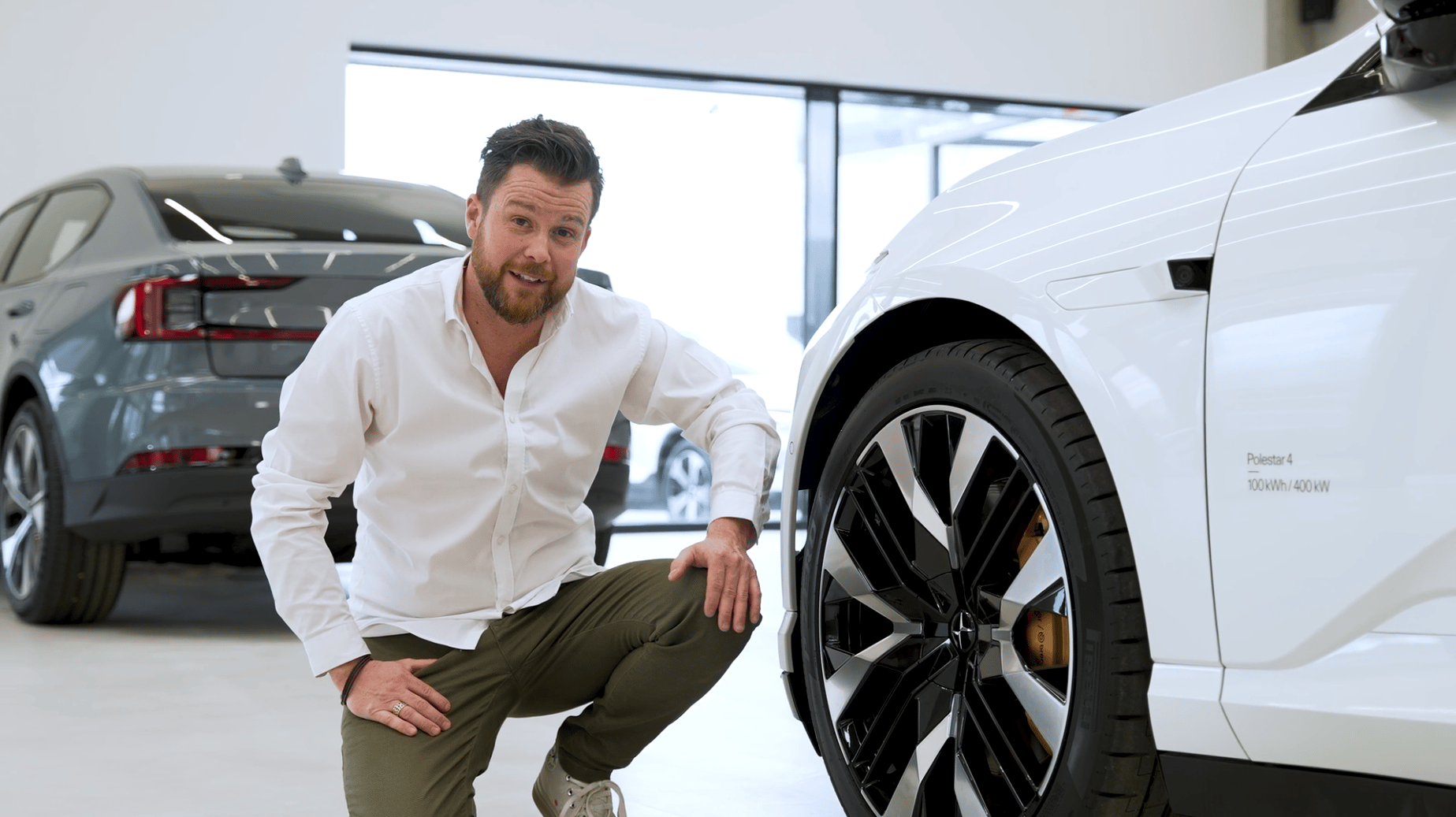 Person crouching near a white Polestar car with visible wheel, another Polestar car in the background inside a showroom.