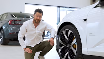 Person crouching near a white Polestar car with visible wheel, another Polestar car in the background inside a showroom.