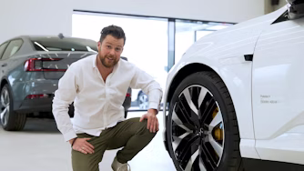 Person crouching near a white Polestar car with visible wheel, another Polestar car in the background inside a showroom.