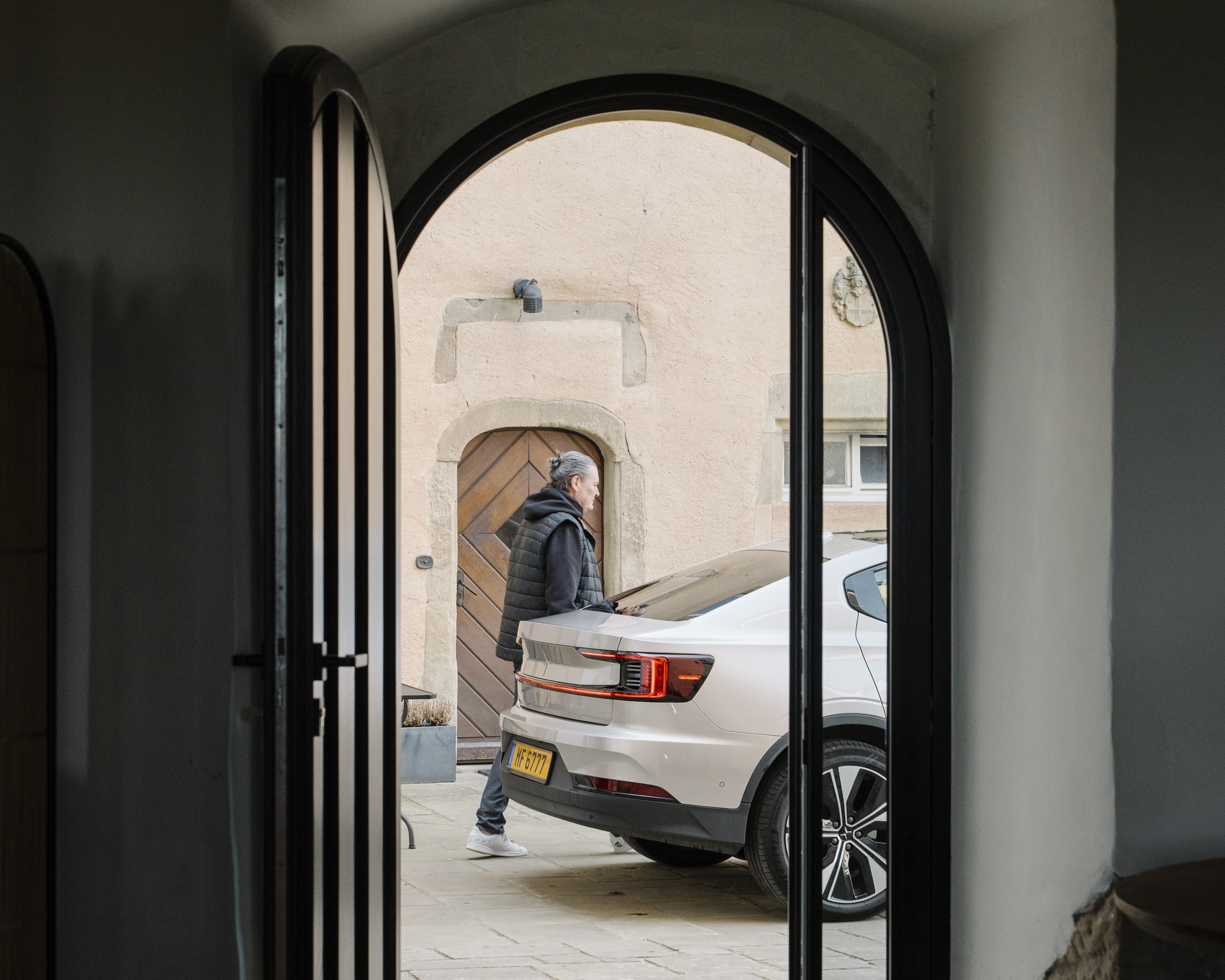 White Polestar car parked near a building with arched doorway, viewed through an open door.