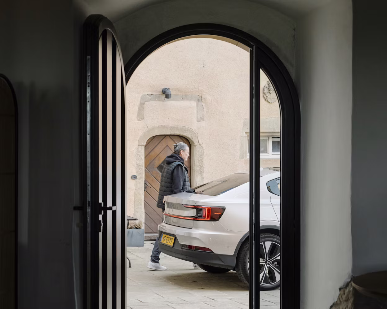 White Polestar car parked near a building with arched doorway, viewed through an open door.