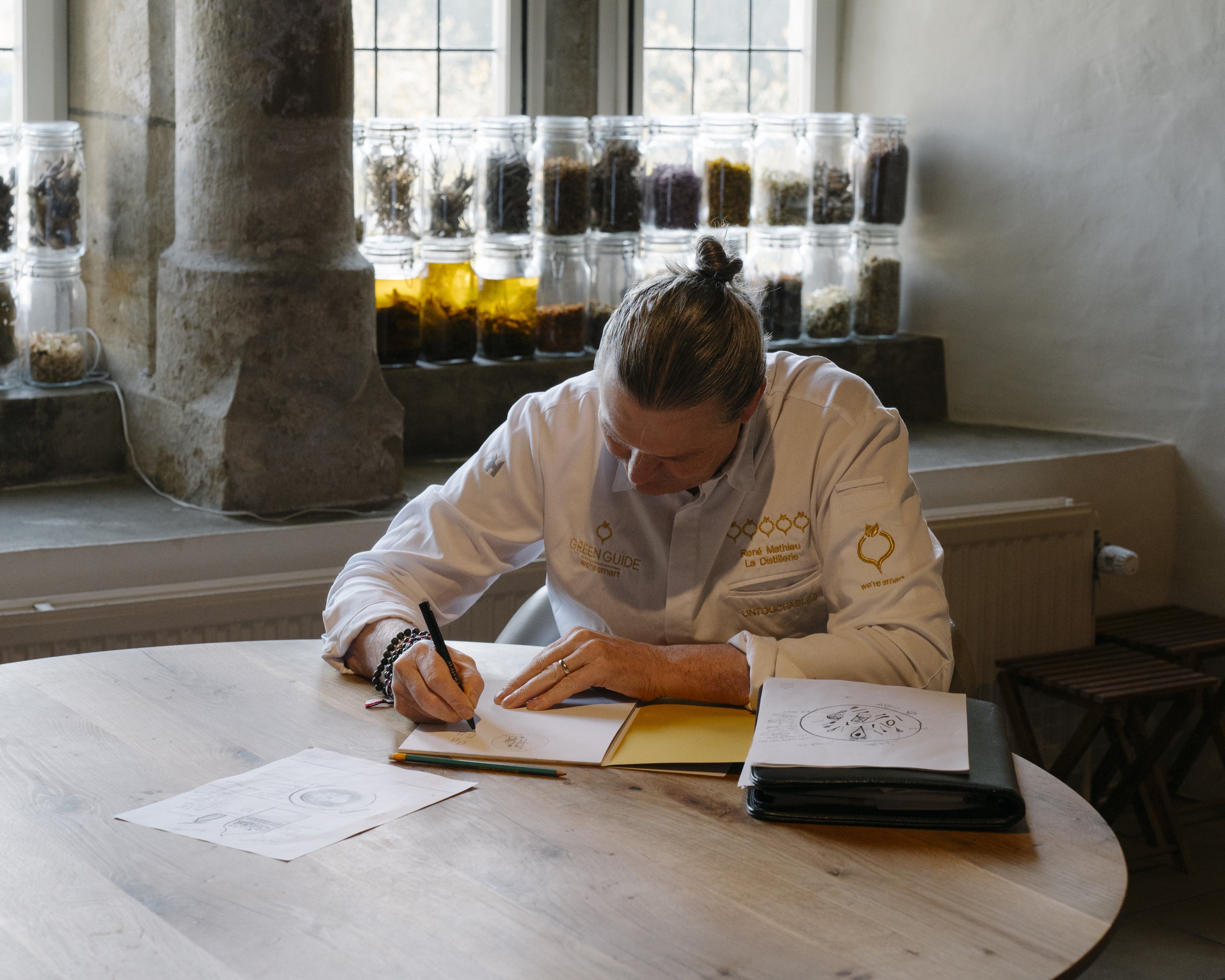 Person in chef coat writing at a table with jars of ingredients on a windowsill in the background.