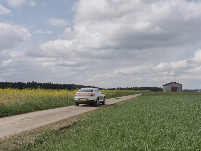 Polestar car on a rural road surrounded by fields and a small building in the distance under a cloudy sky.