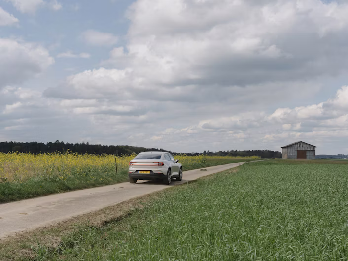 Polestar car on a rural road surrounded by fields and a small building in the distance under a cloudy sky.