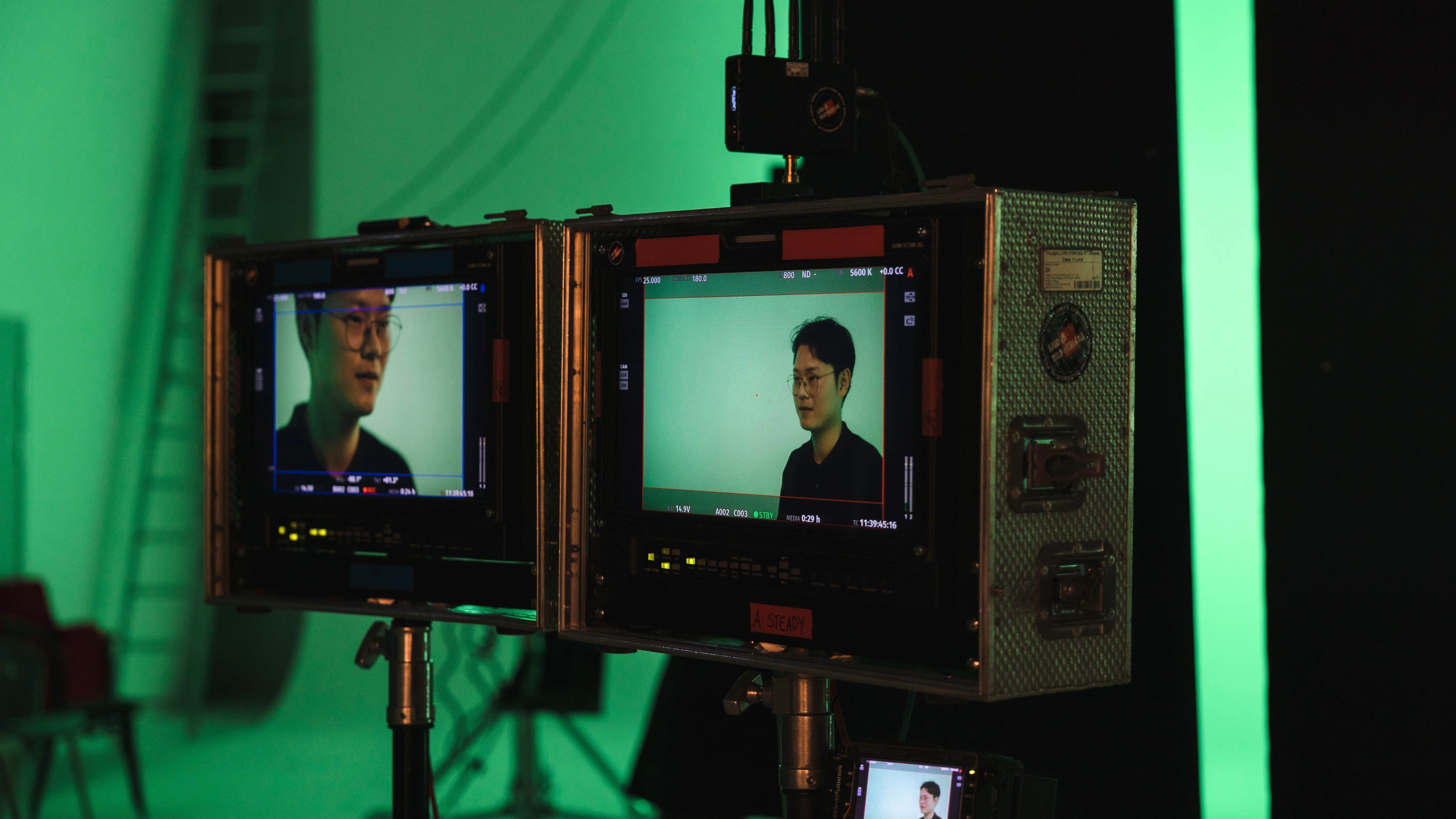 Two monitors display a person on a green screen set with studio equipment and a ladder in the background.