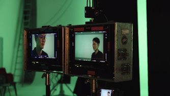 Two monitors display a person on a green screen set with studio equipment and a ladder in the background.