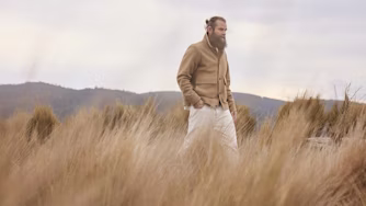 Man walking through tall grass with hills in the background.