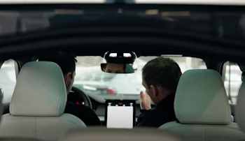 Interior view of Polestar car with two people seated in front, central touchscreen visible, exterior cars in background.