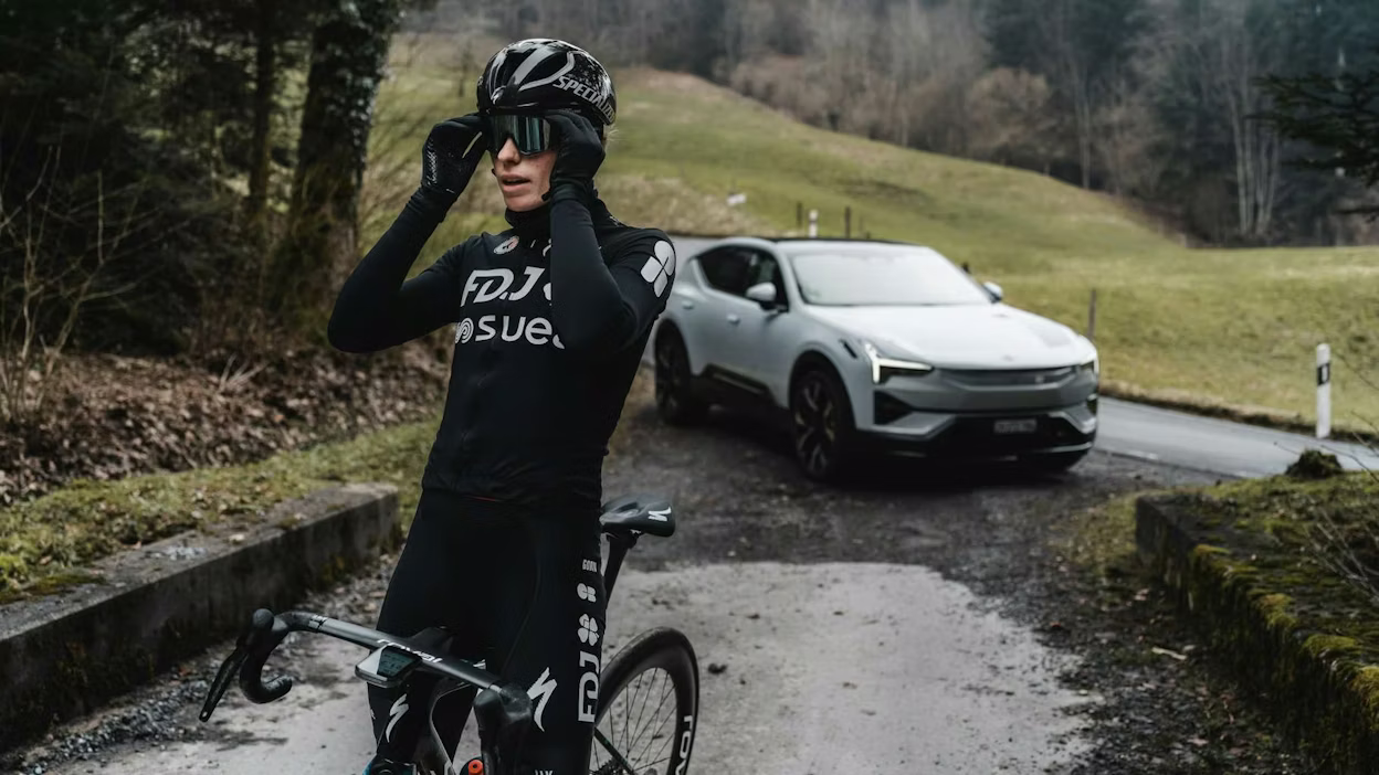 Demi Vollering op een landweg met racefiets, staat voor een witte Polestar 3.