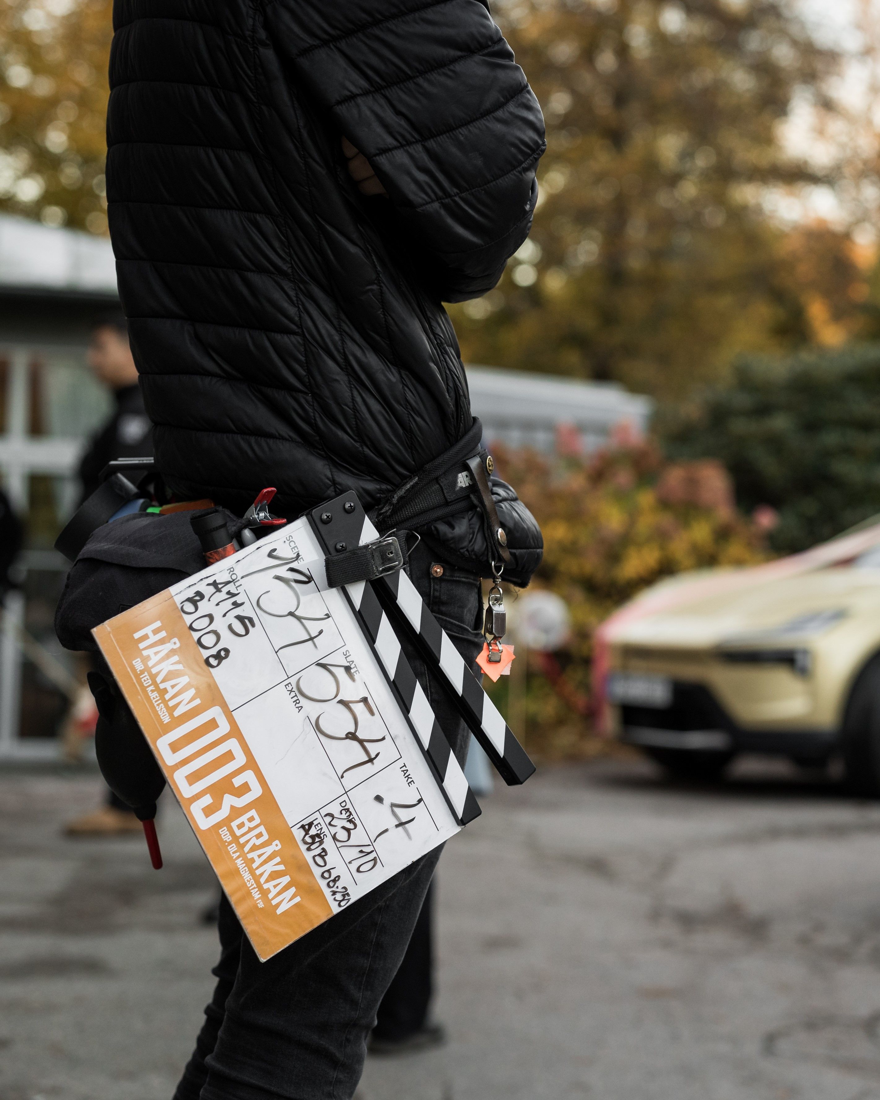 A person standing crossed armes in a black jacket and a movie recording gear in the belt, golden Polestar 4 in the background