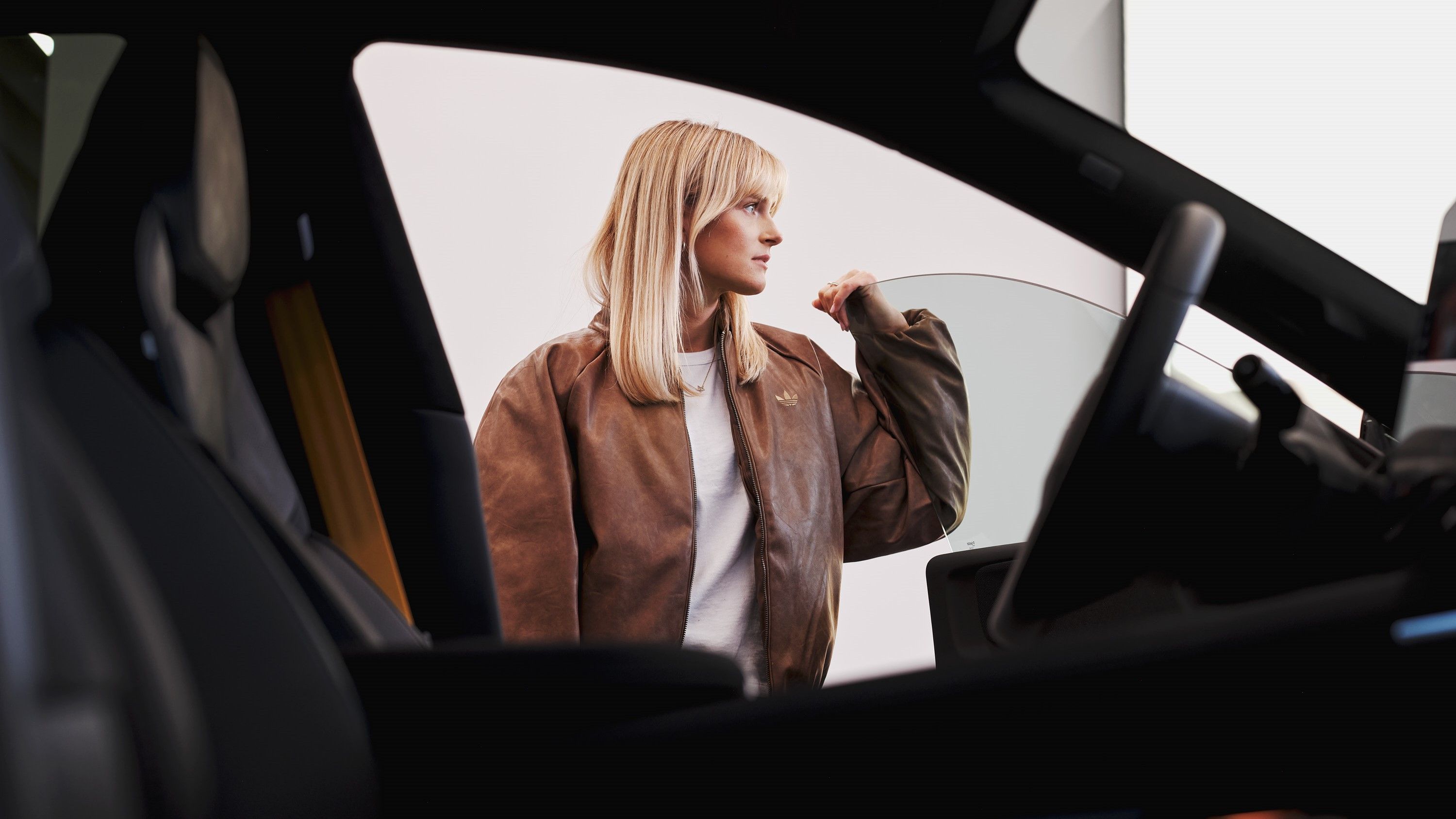 Blonde girl in brown jacket stands outside a car with her hand on the door
