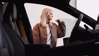 Blonde girl in brown jacket stands outside a car with her hand on the door