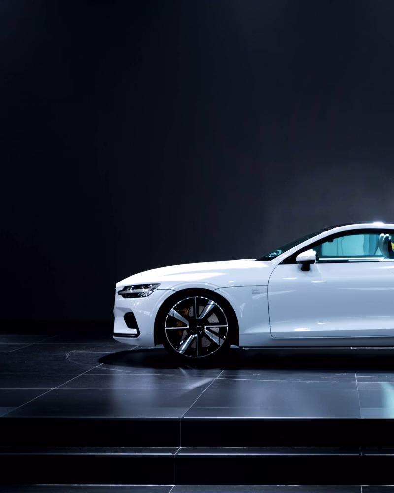 Side view of a white Polestar car on a black platform with dark background.