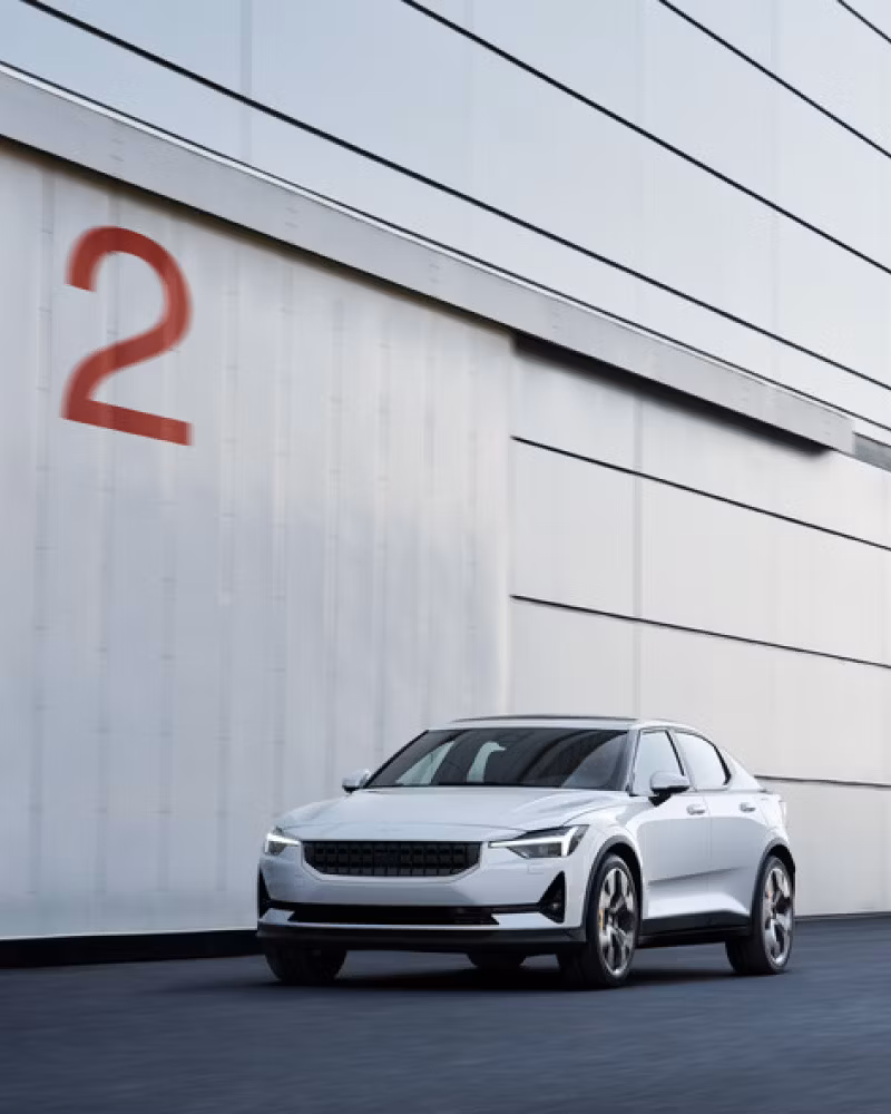 A white Polestar 2 parked infront of a grey garage with a big red 2 painted on the wall