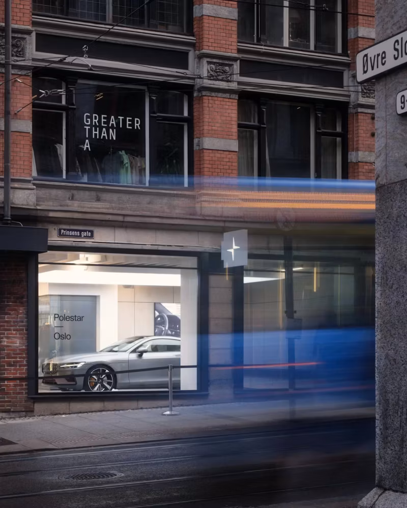 Polestar car displayed in showroom with brick building, tram motion blur, and street signs visible.