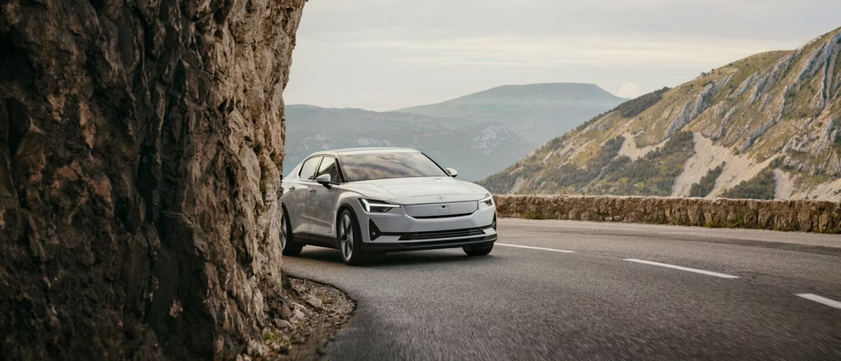 A white Polestar 2 driving around a corner on a mountain road, with rocks to the left and a mountain valley in the background.