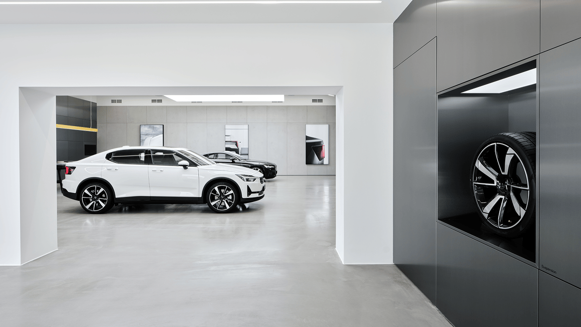 White Polestar 2 car in showroom with another car in background and a wheel displayed in a wall compartment.