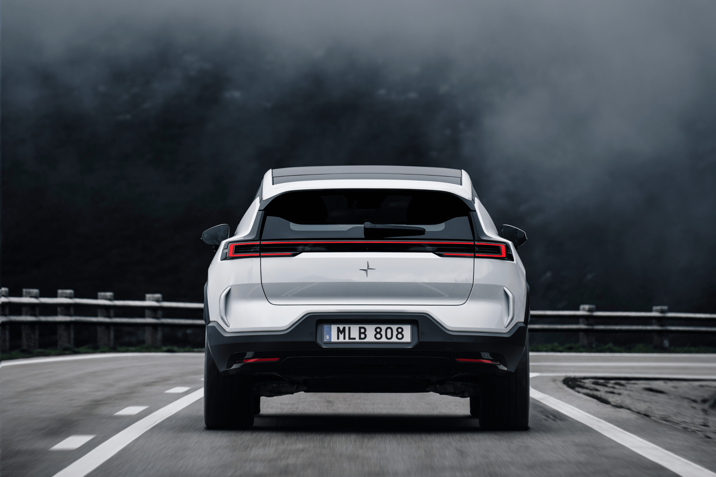 View of a white Polestar 3 from behind driving on a motorway against a foggy, mountainous backdrop.