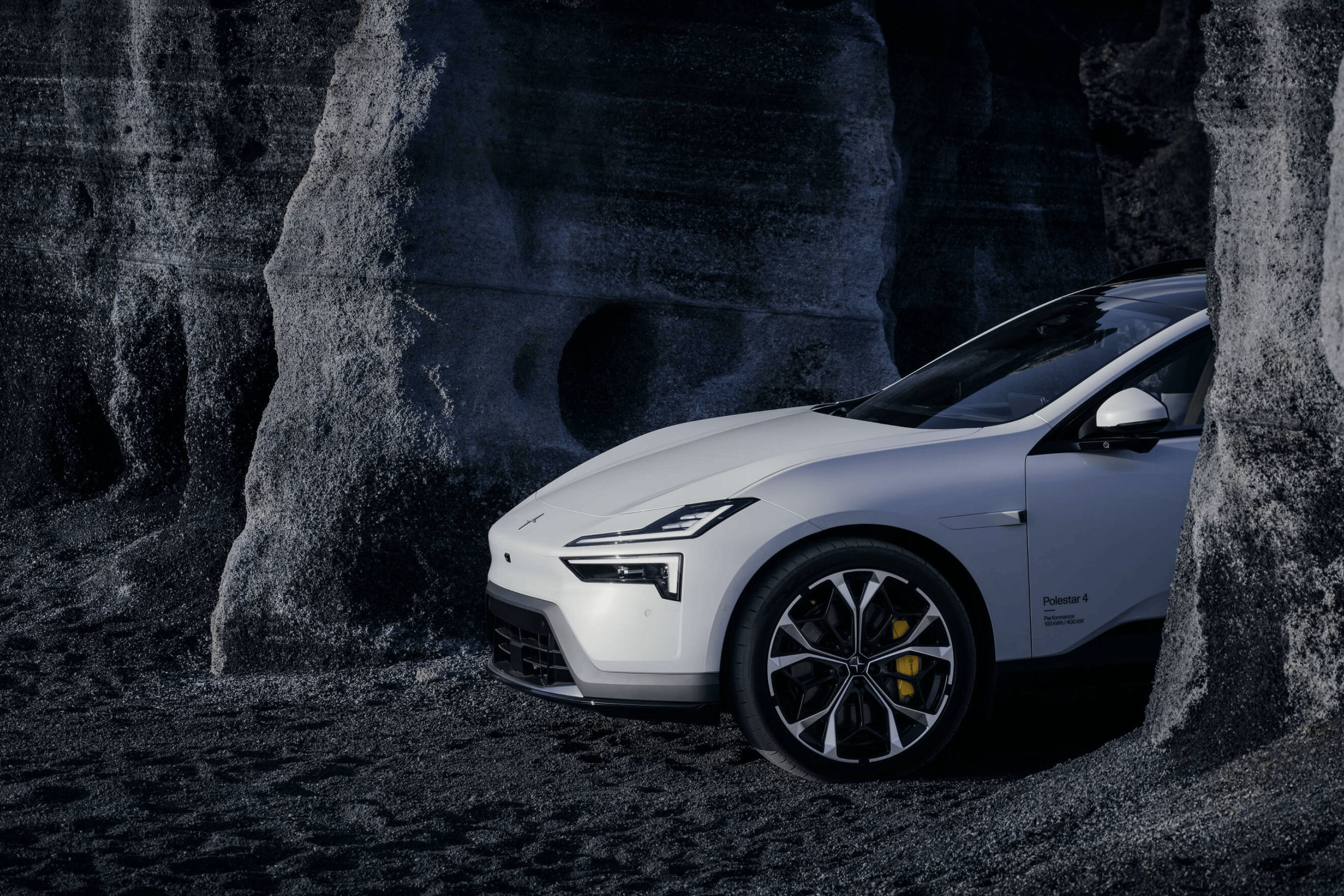 Front side portion of a white Polestar 4 SUV standing between a grey rock formation.