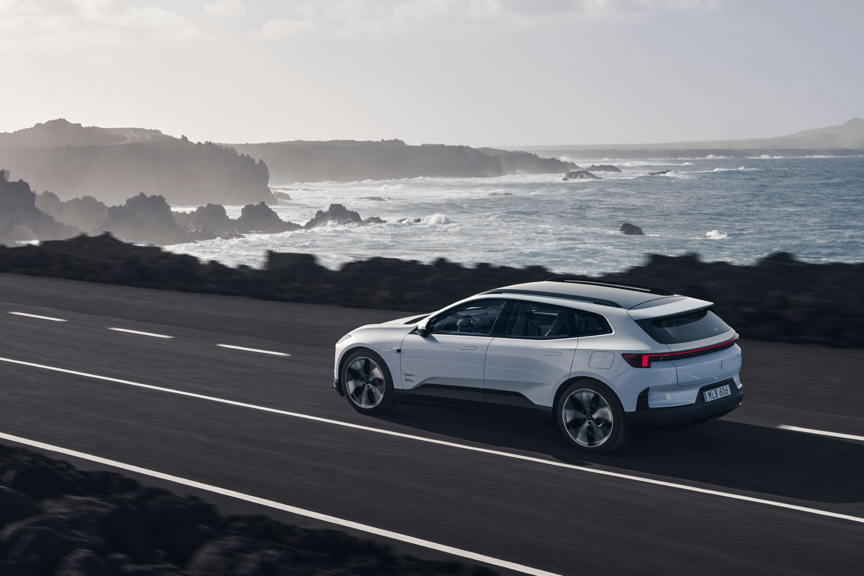 A white Polestar 4 SUV facing left and driving on a dark road by the ocean and rocky sea cliffs.