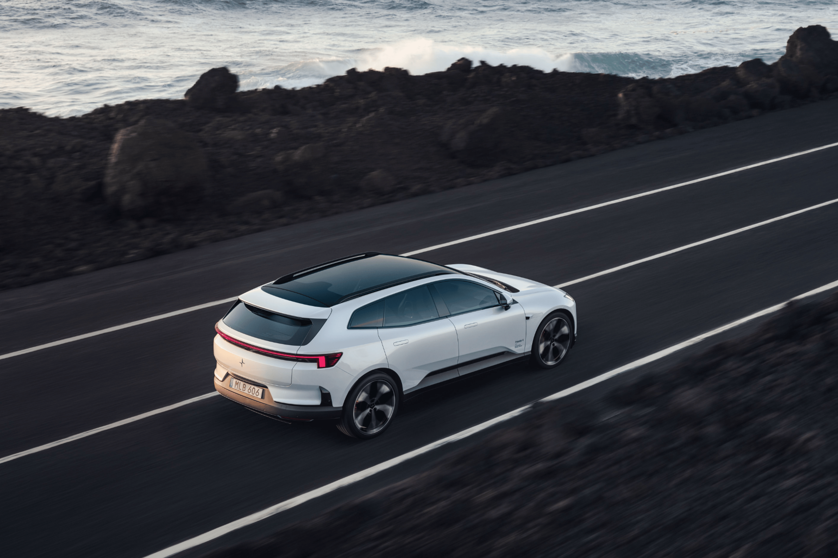 A white Polestar 4 SUV facing right driving on a dark road by the ocean and dark rocks.