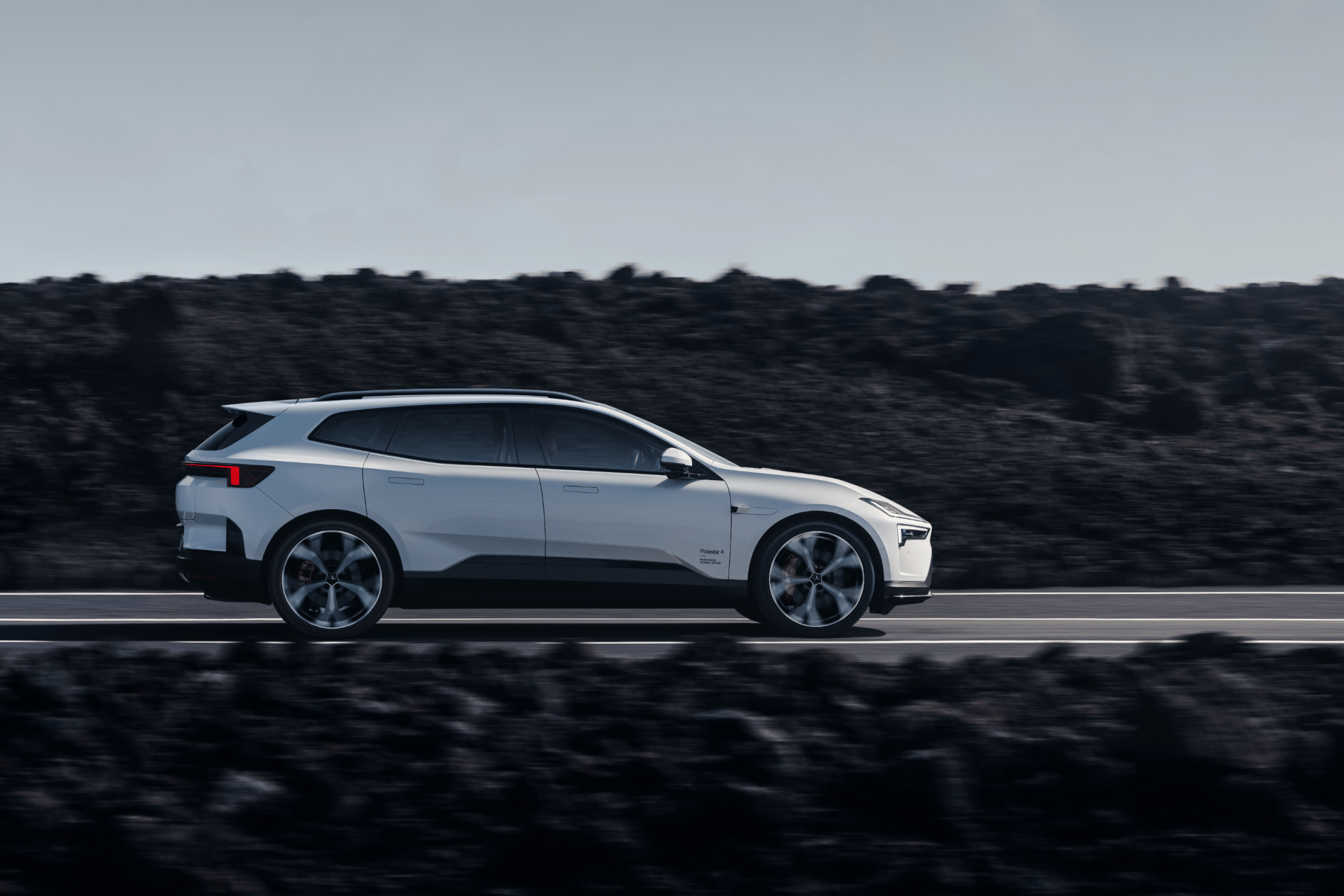 Side profile of a white Polestar 4 SUV driving on a road with dark rocky cliffs in the background.