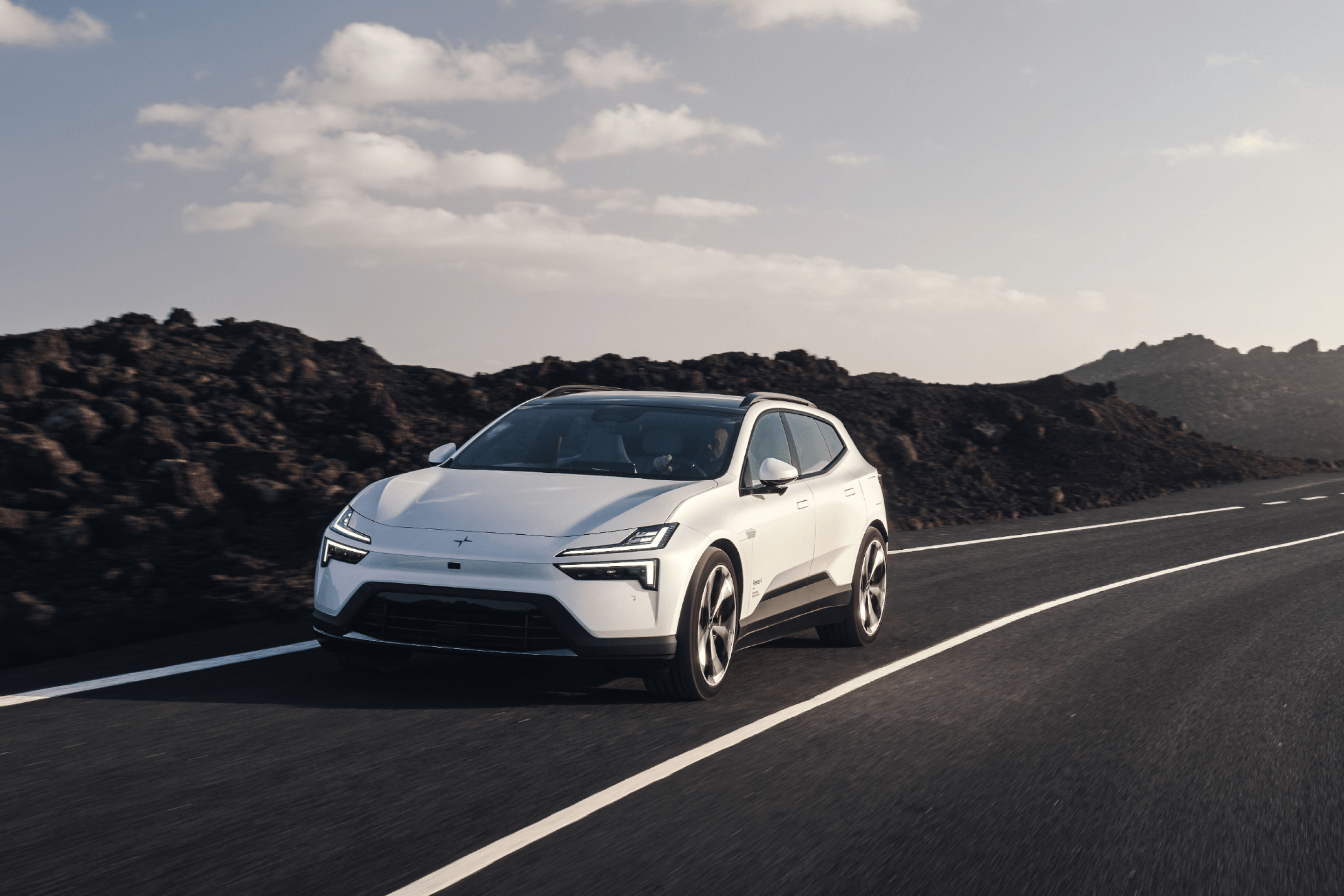Front view of a white Polestar 4 SUV driving down a road surrounded by dark rocky terrain.